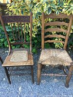 Two vintage wooden chairs standing side by side outdoors on gravel with variegated green and yellow plant background. Chair on left has carved back and caned seat. Chair on right has ladder back and damaged woven seat.