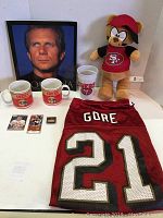 Photo of two white ceramic mugs with San Francisco 49ers logo, a white plastic cup with 49ers and Super Bowl logos, red football jersey with #21 Gore, San Francisco 49ers plush toy, Joe Montana trading card, pin, and ticket stub.