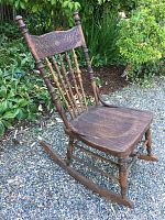 Full view of vintage wooden rocking chair with carved decorative backrest and turned spindles, set on rockers outdoors on gravel with plants behind.