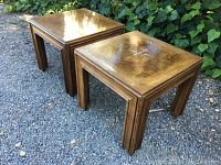 Two square wooden end tables with a warm brown glossy finish, showing wear and abrasions on the top surfaces and legs, set outdoors on gravel with plants in the background.