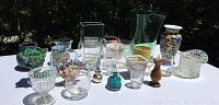 Wide view of 21 assorted vases including clear glass, colored glass, ceramic, and wooden pieces of varying shapes and sizes on white table outdoors