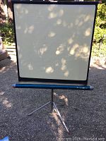 Vintage movie screen fully extended and mounted on tripod stand. Shows off-white screen with black border outdoors under natural light.