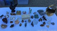 Wide view of assorted figurines, trinket boxes, paperweights, and decorative animals displayed on white cloth showing variety of styles and materials