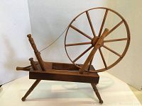 Side view of the antique wooden spinning wheel showing large wheel with spokes, base with three legs, and spindle components.