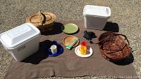Photo showing all picnic items arranged on a brown blanket placed on gravel: two white Styrofoam coolers, two wicker baskets, plastic plates and cups, a small folded umbrella, bamboo plate holder and skewers, vintage green platter.