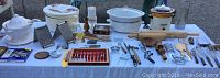 Full table view showing all kitchen cooking items including crockpots, dehydrator, graters, and utensils spread on white tablecloth outdoors.