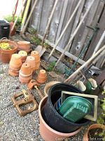 Clay pots, plastic pots, various small gardening containers, and garden hand tools such as a pair of hand forks or clippers arranged on gravel ground near fence.