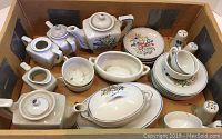 Full overview of children's porcelain tea set pieces inside cardboard box, showing various teapots, cups, saucers, serving dishes, and salt & pepper shakers.