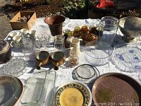 Photo showing a variety of glass, ceramic, and metal tableware laid out on a white tablecloth outdoors, including clear and amber glass cups, jars, ceramic pots, mugs, plates, and a large ornate metal bowl.