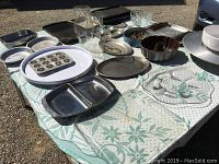 Outdoor photo showing an array of trays, platters, baking tins, bowls, and glasses laid out on a patterned cloth, highlighting the variety and condition of the serving and baking items.