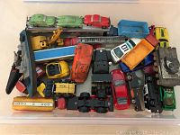 Overhead photo showing the assortment of vintage metal toy cars and trucks in a plastic container. Featured cars include green and red vintage cars, a car carrier truck with cars, a yellow excavator, fire truck with ladder, white police car, and other assorted vehicles.