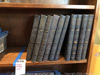 Ten black leather bound books arranged on a wooden shelf. Spines show gold embossed titles and military motifs indicating volumes of Pictorial History of the Second World War, with one additional volume for the Korean War. Volume VI is missing.
