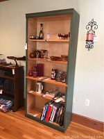 Front view of tall painted wood bookshelf with three shelves displaying various glassware and decorative items. Natural wood tone inside with green-painted exterior edges.
