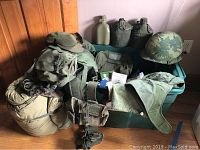 Photo showing lot contents including green sleeping bag, military helmet with camouflage cover, two canteens with canvas covers, assorted bags and belt with pouches arranged on floor.