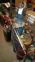 Side view of blue oxygen gas cylinder with regulator and hoses attached, standing next to red acetylene tank and other equipment.