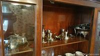 Silver plated tableware displayed on a wood shelf, includes several pieces like salt and pepper shakers, candlesticks, serving bowls, and trays.