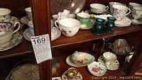 Wide view showing multiple floral patterned fine china teacups, saucers, and other tableware items arranged on wooden shelves.