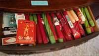 Drawer containing a large assortment of twisted and smooth colored candles in red, green, and cream hues. Includes boxed holiday candles with Christmas branding.