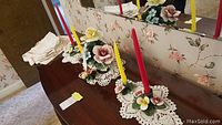 Wide view of Capodimonte porcelain floral candle holders displayed on brown furniture, each holding red and yellow taper candles, accompanied by white crochet-style table linens.