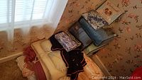 Photo showing assortment of folded and packaged blankets and bed linens in a corner near a window with curtain.