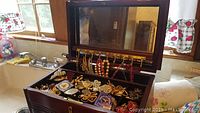 Open wood jewelry box showing assorted costume jewelry inside, earrings hanging inside the lid, various necklaces and pins in compartments.