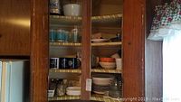 Photo showing open kitchen cupboards with multiple shelves filled with various ceramic mugs, glasses and bowls in different colors and sizes.