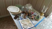 Overview of Bakerite platters, glassware, figurines, silver coaster set and brass hurricane lamp on table