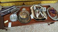 Overview of multiple serving and cooking items displayed on wooden table including silverplated, enamelware trays, utensils, and bowls