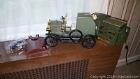 Wide shot of the green vintage die-cast model car decanter alongside a brown leather flask case and a small bagged item, set on a wooden surface.