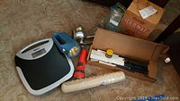 Photo showing assorted items on carpet: digital scale, blue and orange flashlights, vintage metal meat grinder, Ice O Mat ice crusher cardboard box, brown box with carving knives, plastic roll.