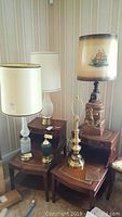 Seven assorted table lamps arranged on wooden tables, showcasing variety of materials including ceramic, brass, and cut glass. Includes the large ships anchor dock lamp with sailboat motif shade visible.