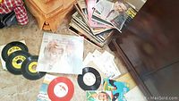 Photo showing a pile of various vintage vinyl records, some 45 rpm singles and full albums, visible sleeve with a female artist photo, some albums stacked on floor near furniture.