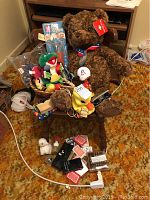 Wide view of the assortment of toys and games on a chair with brown patterned carpet background. Includes Gund Wish Bear plush, Patriots bobblehead, plush duck bank, playing cards, and small boxed games.