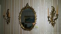 View of oval mirror with decorative brass colored frame flanked by two matching brass colored molded resin candle sconces holding two candles each, mounted on wall with vertical striped wallpaper.