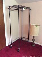 Full view of black metal garment rack with four wheels and brass table lamp with cream lampshade.