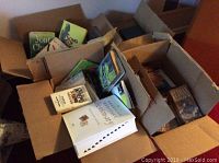 Four open cardboard boxes filled with assorted books including hardcover and softcover novels and reference books.