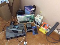 Photo shows the Samsung DVD VHS combo player V2000, boxed Ameritech flip phone, boxed Sony internet terminal for TV, boxed ATT GO phone, surge protector, and several other small electronics items on a floor corner.