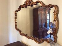 Front view of large wall mirror showing ornate gold frame and reflective surface. Mirror reflects a dining room with wooden furniture.