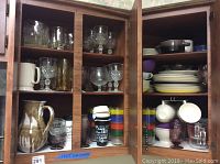 Two kitchen cabinets filled with assorted glassware including clear cut crystal stemware, colored tumblers with stripes, glass bowls, and ceramic and glazed terra cotta pitcher. Lower shelves have assorted everyday dishes, teacups, and soup crocks in various ceramic materials and colors.