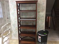 Full view of five shelf wooden storage rack and Rubbermaid trash can positioned to right side of rack in a kitchen corner.