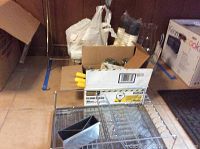 Various candles, glass chimneys, cardboard boxes, wrapping materials, stainless steel dish drainer and tray on tiled floor.