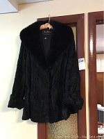 Photo of black lamb fur coat hanging on hanger showing full front of coat and sleeve folds.