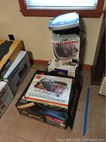 Photo shows a stack of boxed items including a George Foreman Color Grill on top, a Metro roasting/baking/lasagna pan box, and a Maxim wok box at the bottom.