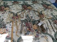 Wide view of assorted necklaces laid on floral fabric background showing various stitches and styles including faux pearls, gold chains and beads.
