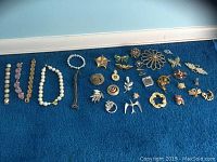Image showing an assortment of pins and brooches in various shapes and designs spread on blue fabric including floral, bird, star, and leaf motifs with mixed metal tones and some rhinestone accents.