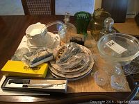 Wide view of lot items including six Corning Ware cookware pieces with glass lid, Fire King casserole dish, multiple glassware items, and silver-plated utensils arranged on table.