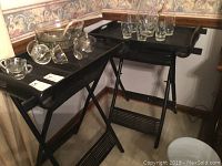 Two black folding tray tables with trays attached, set side by side, with glassware displayed on top of one table.