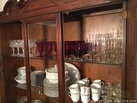 Shelf with various stemware glasses in clear and red and dinnerware including plates, cups, and covered casserole dish within a wooden cabinet