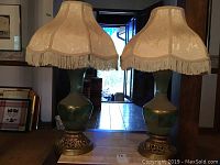 Two vintage lamps with green glass bodies, fringe fabric shades, and ornate gold bases shown together.