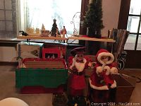 Photo showing two untested animated Christmas figures dressed in red and plaid holding candle-like lights in front of green and red plastic storage bin, with boxes and wrapping paper behind on tables.